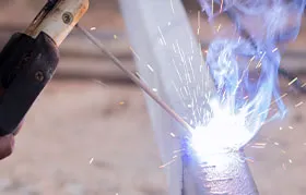 A close-up of a welder performing Shielded Metal Arc Welding (SMAW), or stick welding, showing the intense, bright arc, sparks, and smoke generated as the electrode melts to join the metal parts.