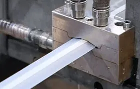 A close-up photograph of an industrial extrusion machine in operation, showing a custom co-extruded plastic or rubber profile emerging from a precision-machined steel die. This demonstrates a real-world manufacturing application.
