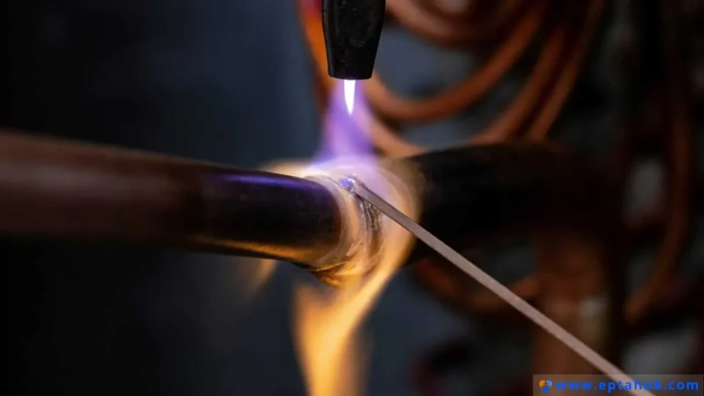 A close-up of torch brazing in action, where a technician uses a hot flame to heat a copper pipe joint and melts a filler rod into the seam, demonstrating the capillary action that forms the bond.