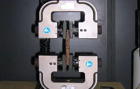 A close-up photograph of a tensile test in progress, with a rectangular composite or wood specimen securely mounted in the heavy-duty mechanical grips of a universal testing machine, ready to be pulled to its fracture point to measure UTS.