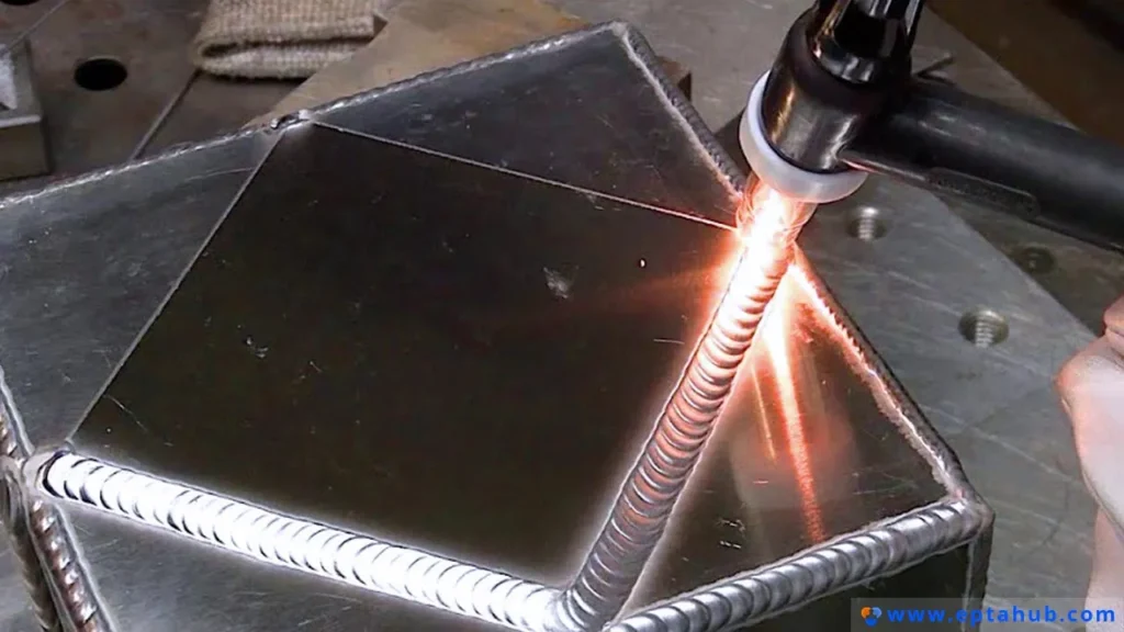Uma imagem em close-up de uma solda TIG de alta qualidade sendo realizada em um cubo de alumínio, mostrando o cordão limpo e preciso, característico do processo GTAW, com aspecto de "pilha de moedas".