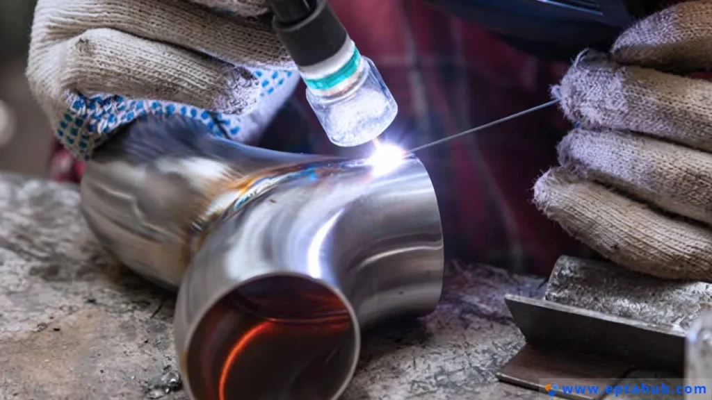 Um close-up de um soldador realizando uma solda TIG de precisão em um cotovelo de tubo de aço inoxidável, alimentando manualmente a vareta de solda para criar uma junta forte e limpa para uma peça personalizada.